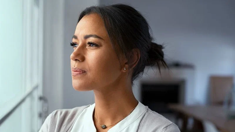 Looking out window TBT Close-up of a woman in a gray sweater, hair pulled back, looking out the window with a thoughtful expression. The blurred modern living room suggests her focus on handling probate real estate mistakes. Contact Kid Breukelen Realty Group for guidance.