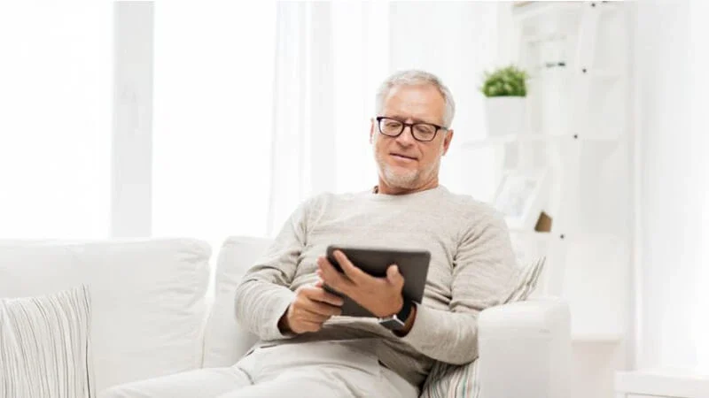 reading tablet TBT Man sitting on a white sofa, reading a tablet in a white-themed room, reviewing essential probate real estate terms. Contact Kid Breukelen Realty Group for assistance.
