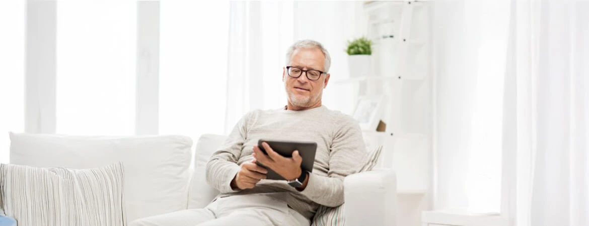 reading tablet DT A man seated on a white sofa in a minimalist white-themed room, reading a tablet. The image illustrates a probate real estate representative reviewing essential terms. Reach out to Kid Breukelen Realty Group for expert help in understanding probate real estate.