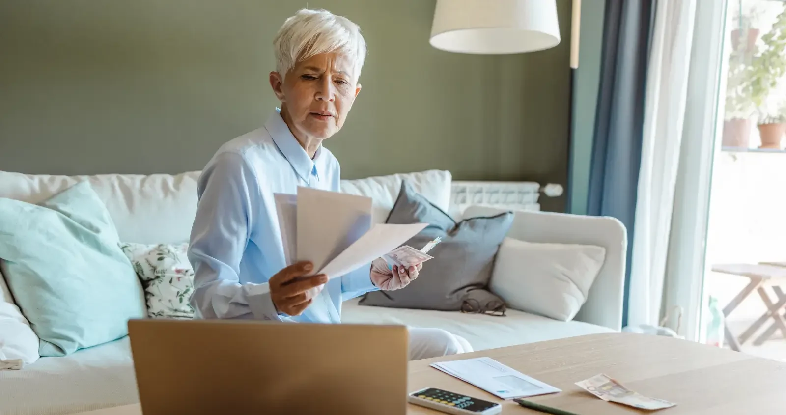 A woman sitting on a couch near a window reviewing bills with a laptop open, representing mortgage or foreclosure-related property stress in 33009.
